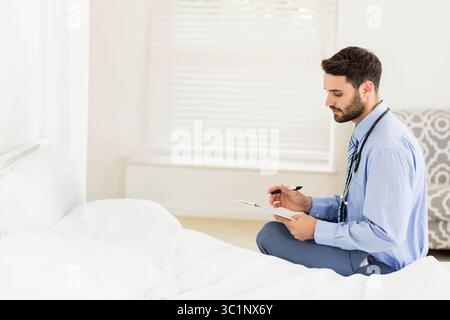 Male doctor in blue shirt holding clipboard while writing notes on bed with stethoscope, copy space Stock Photo