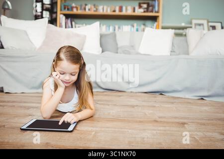 Female child lying on wooden floor in living room tapping tablet near sofa cushions and bookshelf Stock Photo