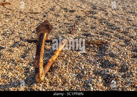 A rusty anchor lying on a shingle beach in warm evening light, weathered by time and tide along the British coast. Stock Photo