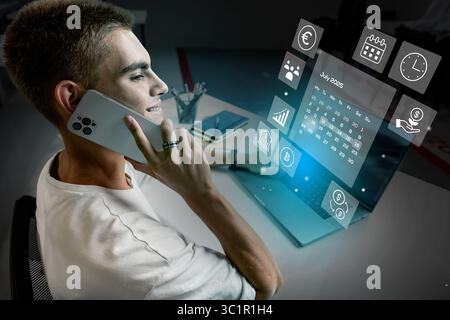 Young professional using smartphone while working on laptop at desk displaying smart technology in modern office space Stock Photo