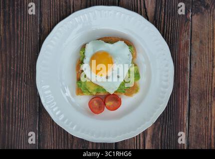 Top view of Heart shaped fried egg on smashed avocado and tomato on a white plate on a wooden background Stock Photo
