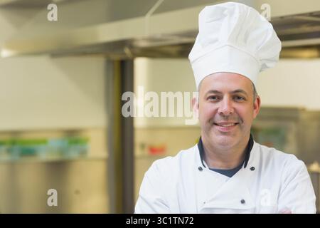 Male chef wearing white chef jacket and hat standing under vent hood in kitchen, copy space Stock Photo
