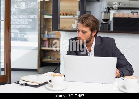 Man wearing dark suit typing on laptop at café table, writing in notebook beside coffee cups Stock Photo
