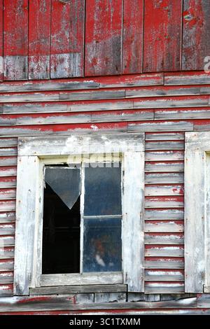 Jagged glass edge hangs from window frame on old, red, wooden barn in Vermont.  Boards on exterior of barn are weathered and very little of red paint Stock Photo