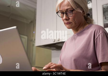A Focused Senior Woman Working on a Laptop in a Modern Kitchen Setting, Emphasizing Technology Usage and Home Office Dynamics Stock Photo