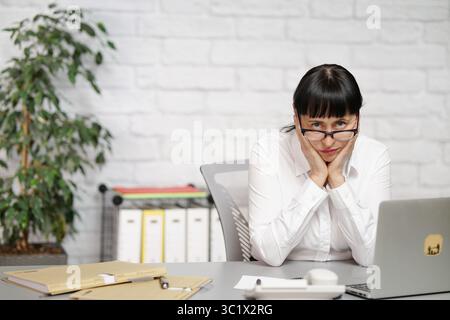 Secretary sitting at desk, head resting on hands, looking at camera, white, mid-age, female, blouse with glasses Stock Photo