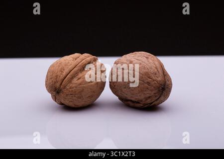 Two walnuts rest side-by-side on a white surface, creating a mirror reflection. The walnuts have rough, textured shells with visible cracks and ridges Stock Photo