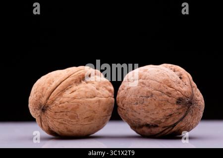 Two walnuts rest side-by-side on a reflective white surface. The walnuts have a rough, textured exterior with deep brown hues and subtle variations in Stock Photo