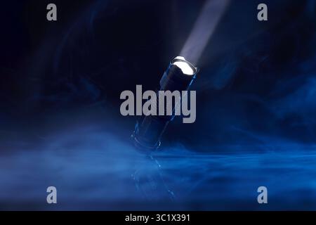 A bright LED flashlight shines through a dense cloud of blue smoke, creating a dramatic beam of light. The flashlight is a metallic silver color The s Stock Photo