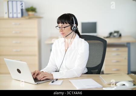 Secretary talking on the phone in the office with a headset and typing on the computer Stock Photo