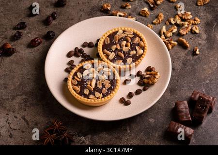 Two dark chocolate tarts sit on a beige plate, garnished with walnuts, coffee beans, and dried cherries. A few chocolate bars are placed beside the pl Stock Photo