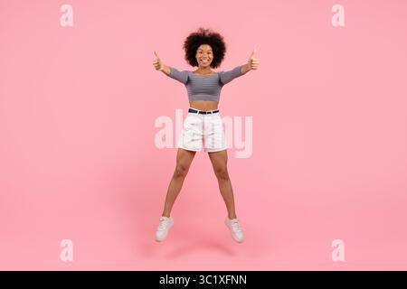 Energetic young black woman jumping up with two thumbs up, smiling confidently Stock Photo