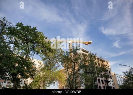 View from below of a construction crane with a building under construction against a blue sky, green trees in the foreground. Stock Photo