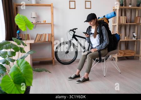 Female hiker traveler with backpack using mobile phone in living room, chats on smartphone app, goes on trip hiking vacation. Copy space Stock Photo