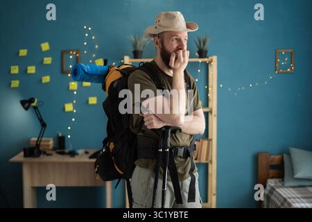 Young bearded man traveler in hat prepare for hiking vacation at home nervous biting nails. Emotion, problem and travel concept Stock Photo