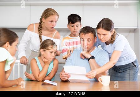 Frustrated man sits in kitchen next to family and reads unpleasant news in letter. Stock Photo