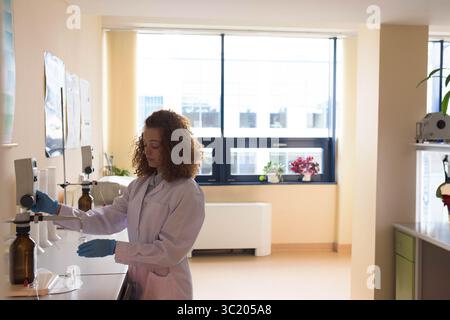 Female lab technician wearing lab coat pouring liquid from amber bottle at lab bench, copy space Stock Photo