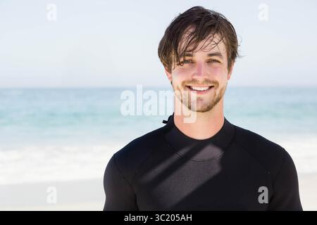 Man standing near waterline on sandy beach wearing black wetsuit and smiling, copy space Stock Photo