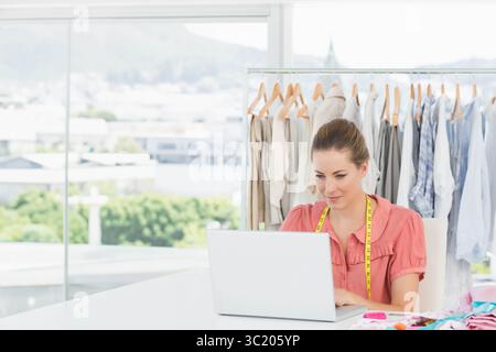 Mid-adult female using laptop wearing measuring tape in fashion studio, clothing rack, copy space Stock Photo