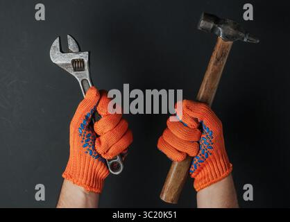 Hands with work gloves are holding a hammer and a wrench on black background. Industrial worker Stock Photo