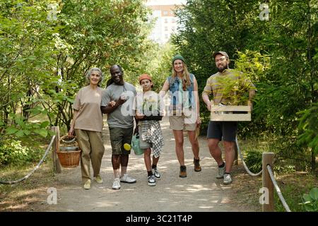Group of diverse adults and one teenager walking outdoors on forest path carrying potted plants and gardening supplies, smiling and looking forward, surrounded by green trees Stock Photo