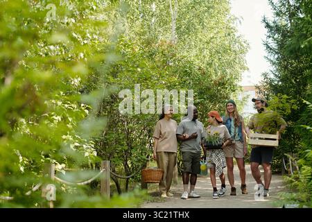 Group of diverse young adults and one senior Caucasian woman walking outdoors on forest path carrying plants and gardening supplies, engaging in conversation and smiling together Stock Photo