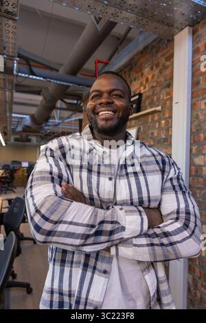 African American man standing with arms crossed and smiling in office with black chairs, ductwork Stock Photo