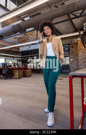 African American woman walking through open plan office talking on smartphone near red-framed desks Stock Photo
