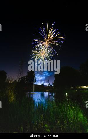 Fireworks burst in vibrant colors over a calm pond, reflecting the dazzling display under a dark sky at night. Stock Photo