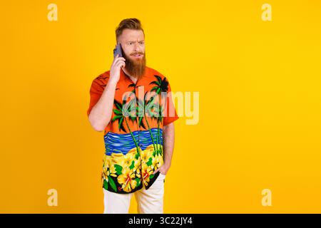 Young bearded man in colorful tropical shirt speaking on mobile phone against bright yellow background, relaxed mood Stock Photo