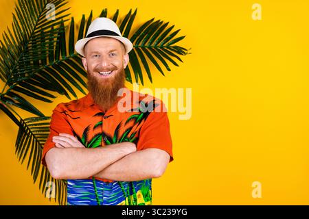 Bearded red-haired man in colorful tropical shirt posing with arms crossed against yellow background with palm leaves Stock Photo