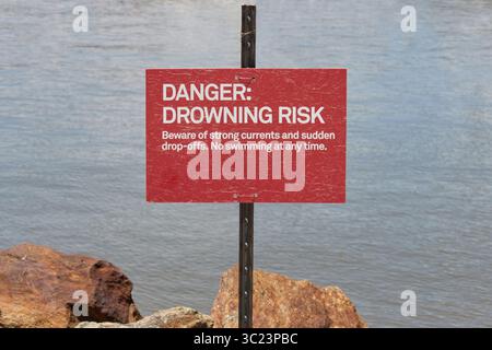 red warning sign with danger, drowning risk, beware of strong currents and sudden drop-offs. No swimming anytime. On a rocky body of water, copy space Stock Photo