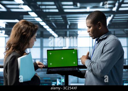 Team of engineers in data center using green screen laptop to do network virtualization. Coworkers in server farm using chroma key notebook, updating storage arrays, verifying settings Stock Photo