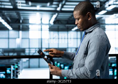 Admin in data center using tablet to deploy new server architectures, configuring network virtualization. Man in server room updating storage arrays, verifying settings to maintain stable performance Stock Photo