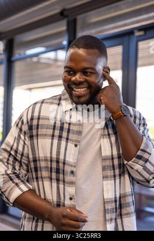 African American man standing at office windows with blinds, touching ear, wearing plaid shirt Stock Photo
