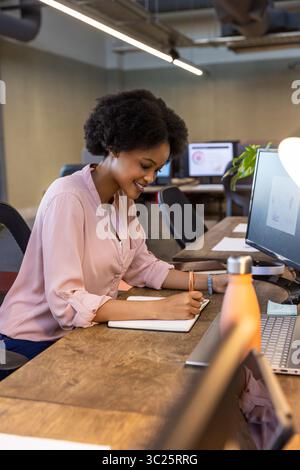 African American woman writing in notebook on office desk in pink blouse with monitors, copy space Stock Photo