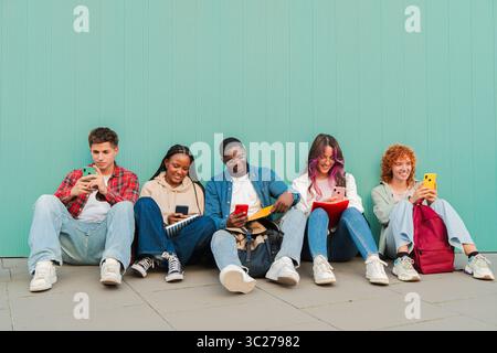 Cheerful group of young friends interacting with their mobile devices, sharing moments and laughter while sitting on the ground, showcasing a modern Stock Photo