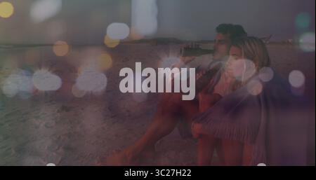Sipping couple wrapped in fringed throw blanket on beach at dusk, with glass bottles, copy space Stock Photo