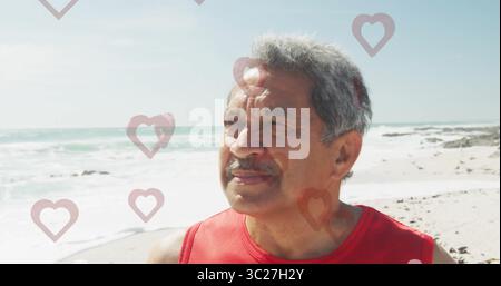 Standing senior wearing red sleeveless top facing ocean on beach, with heart overlays, copy space Stock Photo