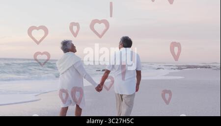 Walking senior couple holding hands along beach at dusk on wet sand, with heart-shaped graphics Stock Photo