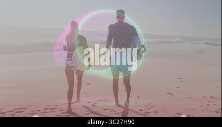 Walking two surfers wearing wetsuits and board shorts on beach, with surfboards and footprints Stock Photo
