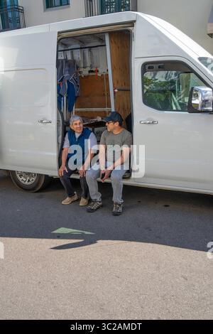 Two generations of men talk while sitting on the floor of an open van door opening during the Tuesday Farmer's Market on State Street in Santa Barbara. Stock Photo
