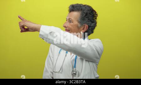 Senior woman doctor with grey hair in white coat pointing against yellow background, holding stethoscope, displaying confidence and professionalism. Stock Photo