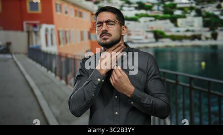 Young man in glasses and gray shirt stands on promenade near seaside holding throat with distressed expression against blurred beach backdrop Stock Photo