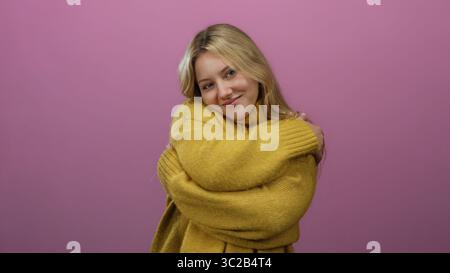Woman hugging herself contently on isolated pink background, wearing a warm yellow sweater with a serene expression embracing self-care and positivity Stock Photo