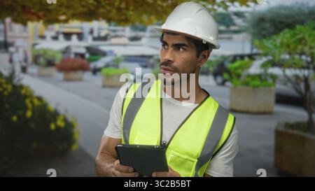 Young hispanic man wearing white hardhat and neon vest holding tablet on busy street; concentration. Stock Photo