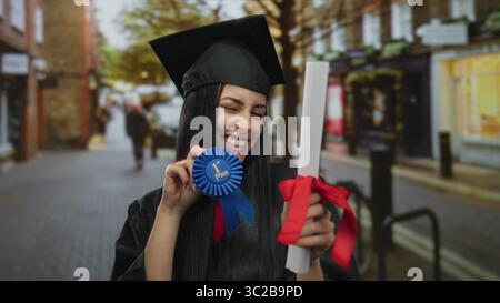 Young hispanic woman in graduation cap and gown proudly holds diploma and blue ribbon while standing on urban street. Stock Photo