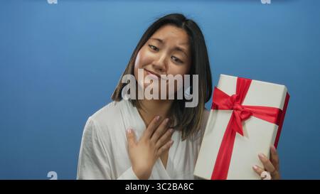 Young chinese woman holding gift box with red bow over blue background, expressing surprise and gratitude. Stock Photo