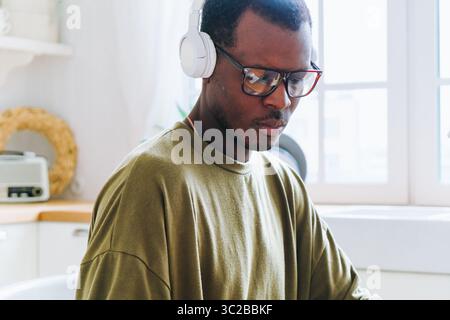 Professional african american male focusing on laptop screen, wearing noise-canceling headphones and stylish eyewear in minimalist home workspace, representing contemporary remote work environment Stock Photo
