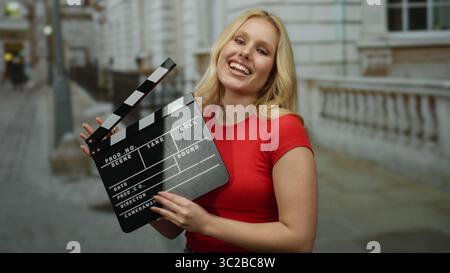 Woman holding clapperboard wearing red shirt smiling on city street outdoors showing filmmaking process. Stock Photo